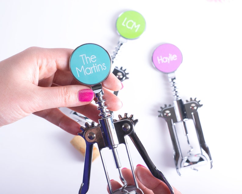 May include: Silver wine bottle opener with a teal circle that says "The Martins". Two other openers are visible, one with "LCM" in green and another with "Haylie" in pink. The openers have a corkscrew design and are held by a person with pink nail polish.