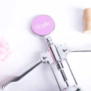 May include: A chrome wine opener with a light purple circular top. The top has the name "Haylie" in white lettering. A cork and pink flower are also in the image.