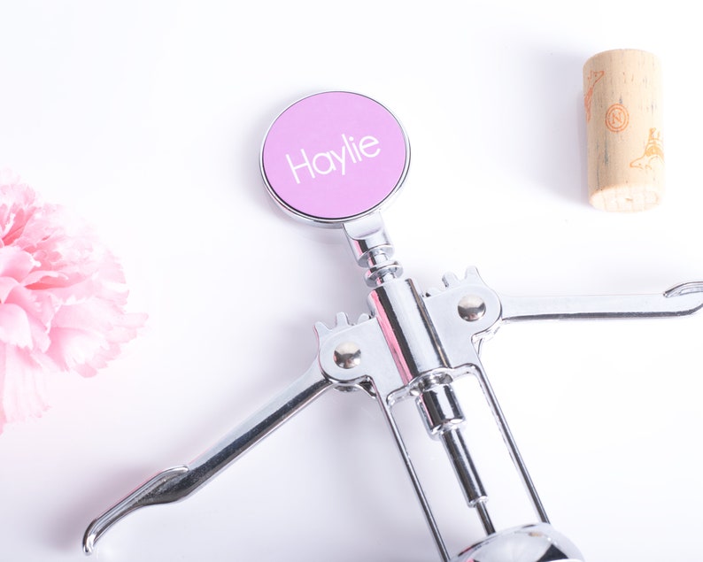 May include: A chrome wine opener with a light purple circular top. The top has the name "Haylie" in white lettering. A cork and pink flower are also in the image.