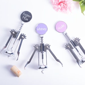 May include: Three personalized wine bottle openers with silver metal bodies and colorful circular tops. The tops are black, lavender, and pink, each with a name printed in white. A cork is also present.
