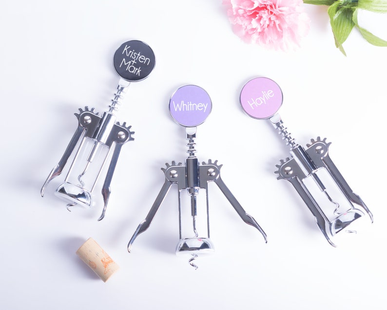 May include: Three personalized wine bottle openers with silver metal bodies and colorful circular tops. The tops are black, lavender, and pink, each with a name printed in white. A cork is also present.