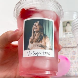 May include: A clear plastic cold cup filled with a pink beverage and ice cubes. A rectangular sticker on the front features a photograph of a smiling person with long hair, with the black handwritten-style text Vintage 1996 below the image.