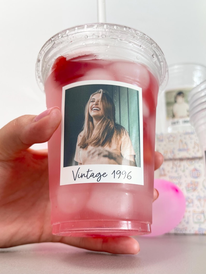 May include: A clear plastic cold cup filled with a pink beverage and ice cubes. A rectangular sticker on the front features a photograph of a smiling person with long hair, with the black handwritten-style text Vintage 1996 below the image.