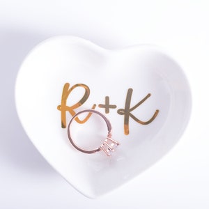May include: White ceramic heart-shaped ring dish with gold lettering that says "R+K". A gold ring with a clear stone is sitting in the dish.