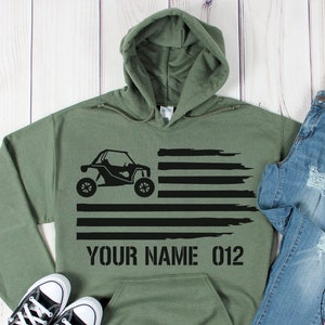 May include: Olive green hoodie with a black graphic of an off-road vehicle and the text "YOUR NAME 012". The hoodie is paired with blue jeans and black and white sneakers. The Cactus Moon Apparel logo is visible.