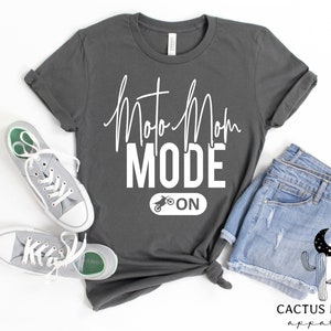 May include: A charcoal grey t-shirt with the text "Moto Mom Mode On" in white lettering. The text "Mode" is in bold, and the text "On" is accompanied by a small graphic of a dirt bike.