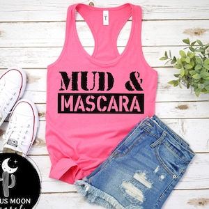 May include: Pink tank top with the text "Mud & Mascara" in black lettering. The tank top is laying on a white wooden surface with a pair of white sneakers and a pair of blue denim shorts.