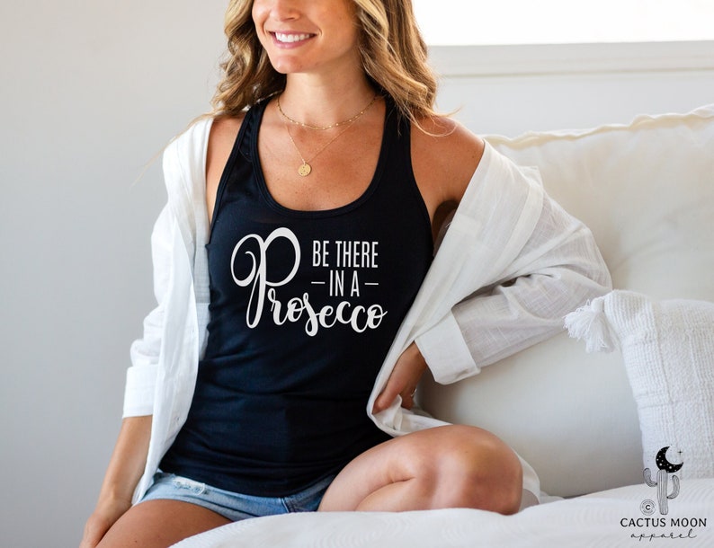 May include: A black tank top with the text "Be there in a Prosecco" in white lettering. The tank top is being worn by a person sitting on a white couch.