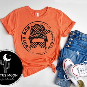 May include: Orange t-shirt with a black graphic of a woman with a checkered bandana and sunglasses. The graphic is in a circle with the text "Moto Mom" and "#dirtbikelife".