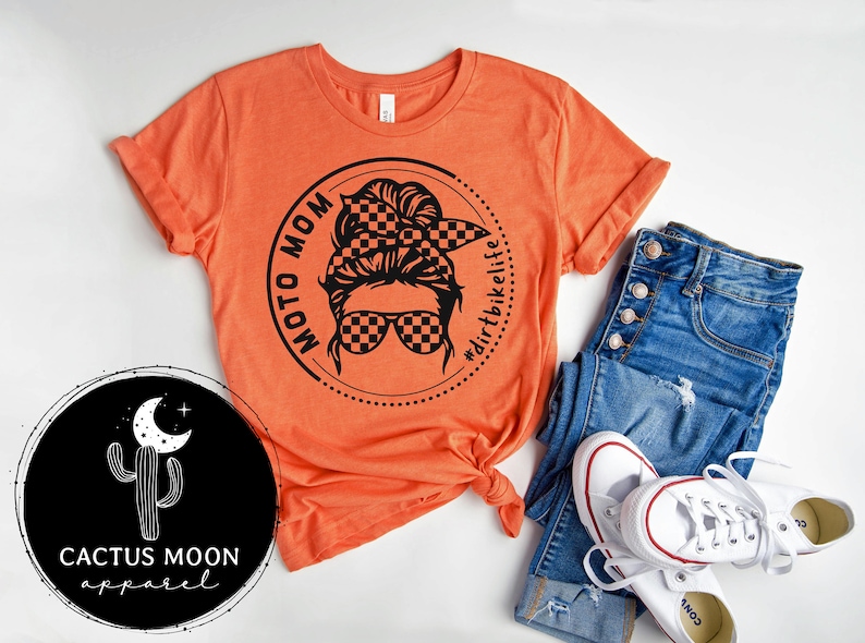 May include: Orange t-shirt with a black graphic of a woman with a checkered bandana and sunglasses. The graphic is in a circle with the text "Moto Mom" and "#dirtbikelife".