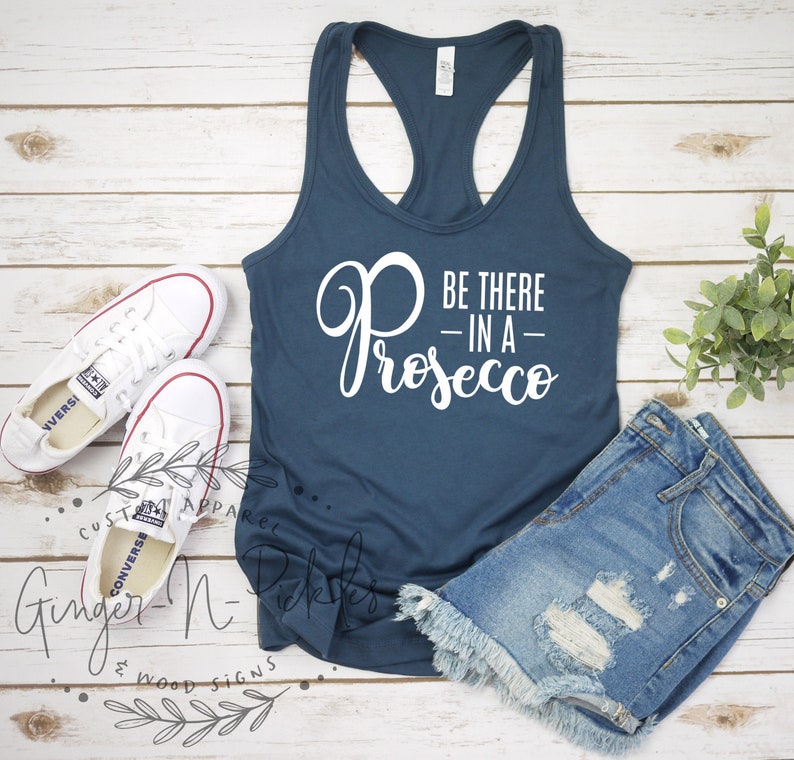 May include: A navy blue tank top with the text "Be there in a Prosecco" in white lettering. The tank top is laying on a white wooden surface with a pair of white sneakers and a pair of blue denim shorts.