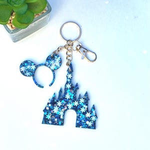 May include: A blue glitter keychain featuring a castle and mouse ears design. The castle and ears are filled with blue and white glitter and snowflake accents. The keychain has a gold-colored clasp and key ring.