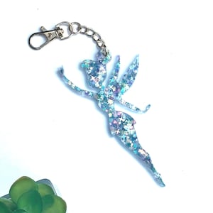 Puede incluir: Un llavero de hada azul brillante con una cadena y un cierre plateados. El hada tiene los brazos y las alas extendidos, con una falda y un diseño en forma de estrella. El llavero está sobre un fondo blanco.