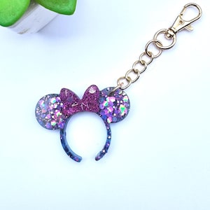 May include: A purple and blue glitter mouse ear headband keyring with a gold-coloured clasp and chain. The headband has a bow in the centre. The ears and headband are filled with glitter.