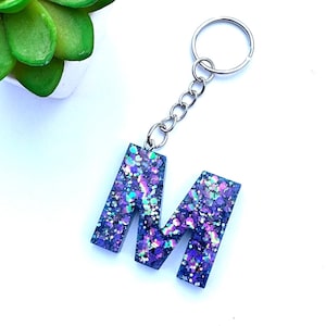 Puede incluir: Un llavero con la letra "M" en purpurina morada con una cadena plateada. La letra de resina está rellena de purpurina iridiscente en tonos morados, azules y verdes. Un accesorio personalizado y decorativo.