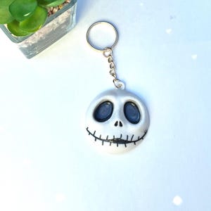 May include: A white Jack Skellington keyring with a silver chain. The round pendant features Jack's face with black eyes and a stitched smile. The keyring is a decorative accessory.