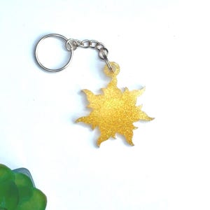 Puede incluir: Un llavero dorado con forma de sol brillante. El llavero tiene una cadena y un llavero plateados. El diseño del sol tiene rayos puntiagudos y un acabado brillante. El fondo es blanco.