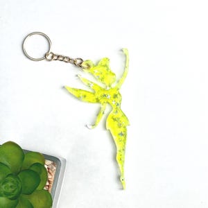 Puede incluir: Un llavero amarillo con forma de hada con detalles de estrellas plateadas. El llavero tiene un anillo y una cadena dorados. El hada está posada con un brazo levantado y una pierna extendida. Una pequeña planta suculenta está en la esquina inferior izquierda.
