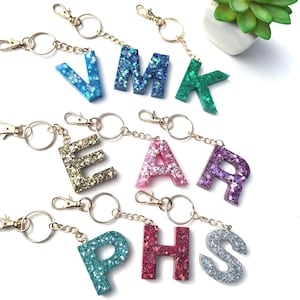 May include: Glitter resin letter keychains in various colors, including blue, green, gold, pink, purple, and silver. Each letter is attached to a gold-tone chain and clasp. The letters are V, M, K, E, A, R, P, H, and S.