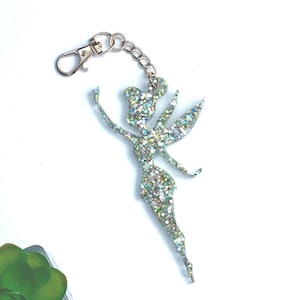 Könnte beinhalten: Glitzer-Feen-Schlüsselanhänger mit silbernem Karabiner und Kette. Die Fee besteht aus schillerndem Glitzer, mit ausgestreckten Armen und detaillierten Flügeln. Ein skurriles Accessoire.