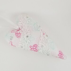 May include: A heart-shaped ornament with a white ribbon for hanging. The ornament is white with a pink floral pattern and white lace trim. Small clear rhinestones are scattered across the surface.