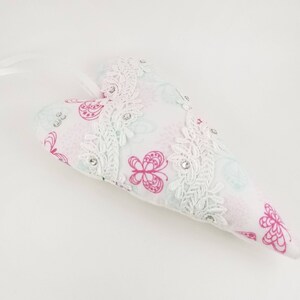May include: Heart-shaped decorative pillow with a white lace trim and pink butterfly pattern. The pillow has a white ribbon for hanging and is embellished with small, sparkling rhinestones.