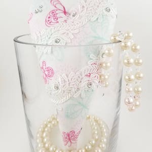May include: A heart-shaped fabric ornament with a butterfly pattern in pink and teal, adorned with white lace and rhinestones, sits in a clear glass vase. A strand of faux pearls encircles the base and drapes over the side.