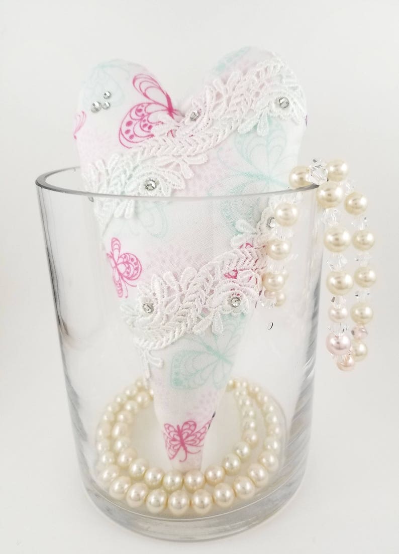 May include: A heart-shaped fabric ornament with a butterfly pattern in pink and teal, adorned with white lace and rhinestones, sits in a clear glass vase. A strand of faux pearls encircles the base and drapes over the side.