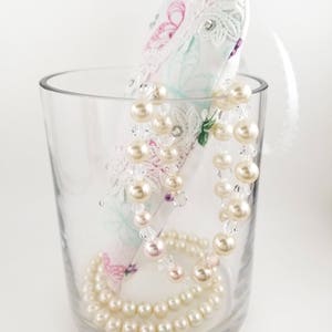 May include: A clear glass vase holds a decorative, white, heart-shaped item with lace and floral patterns. A strand of faux pearls and clear beads is draped over the heart and inside the vase. A second strand of pearls rests at the bottom.