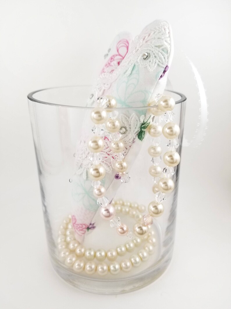 May include: A clear glass vase holds a decorative, white, heart-shaped item with lace and floral patterns. A strand of faux pearls and clear beads is draped over the heart and inside the vase. A second strand of pearls rests at the bottom.