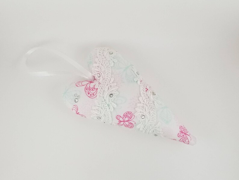 May include: A heart-shaped ornament with a white ribbon for hanging. The ornament is white with a pink floral pattern and white lace trim. Small clear rhinestones are scattered across the surface.