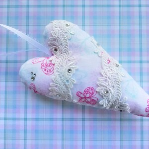 May include: A heart-shaped ornament with a white ribbon for hanging. The ornament is white with pink butterfly patterns and embellished with white lace and rhinestones. The background is a plaid pattern in shades of blue and pink.