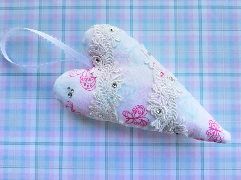 May include: A heart-shaped ornament with a white ribbon for hanging. The ornament is white with pink butterfly patterns and embellished with white lace and rhinestones. The background is a plaid pattern in shades of blue and pink.