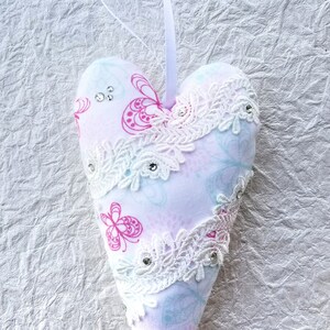 May include: A heart-shaped ornament with a white ribbon for hanging. The ornament is made of fabric with a pink butterfly pattern and white lace trim. Embellished with small rhinestones, it is a decorative item.