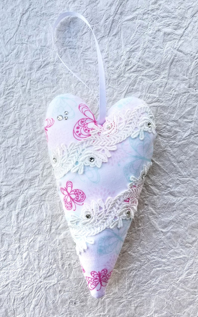 May include: A heart-shaped ornament with a white ribbon for hanging. The ornament is made of fabric with a pink butterfly pattern and white lace trim. Embellished with small rhinestones, it is a decorative item.