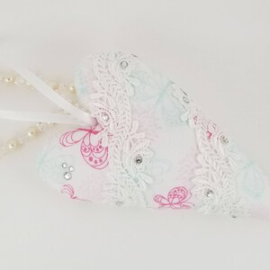 May include: A heart-shaped ornament with a white background. The ornament is decorated with pink butterfly patterns, white lace trim, and small rhinestones. A string of pearls and a white ribbon are attached.