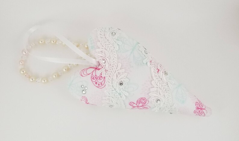 May include: A heart-shaped ornament with a white background. The ornament is decorated with pink butterfly patterns, white lace trim, and small rhinestones. A string of pearls and a white ribbon are attached.