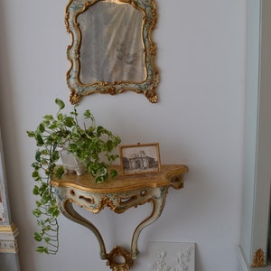 May include: A white and gold ornate console table with a carved design. The table has a mirror with a gold frame and floral details above it. A potted plant sits on the table, and a framed black and white photo is on the right side of the table.