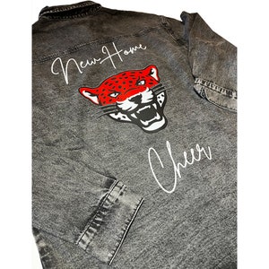 May include: A gray denim jacket with a red and black jaguar graphic on the back. The text "New Home Cheer" is written in white on the back of the jacket.