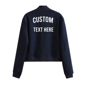 May include: A navy blue bomber jacket with a white embroidered design on the back that says "CUSTOM TEXT HERE".