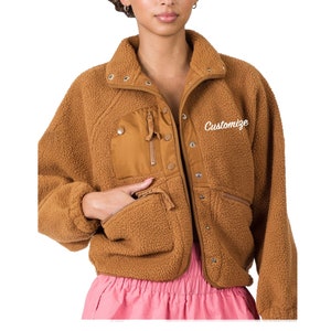 Op de afbeelding: Een bruin fleecejack met een rits en drukknoopsluiting. Het jack heeft een grote zak op de linkerborst en twee kleinere zakken aan de onderkant. Het jack heeft het woord "Customize" geborduurd op de rechterborst.