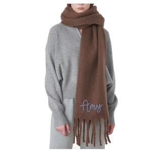 May include: A brown scarf with fringe and the name "Amy" embroidered in light blue. The scarf is wrapped around the neck and shoulders. The model is wearing a gray sweater and pants.