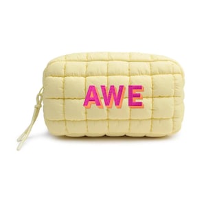 May include: A light yellow, quilted pouch with the word "AWE" embroidered in pink and orange. The pouch has a rectangular shape with rounded edges and a small loop on the side.