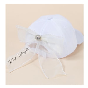 May include: White baseball cap with a large white bow and a rhinestone embellishment. The bow has a long tail with the text "Mrs. Wright" written on it.