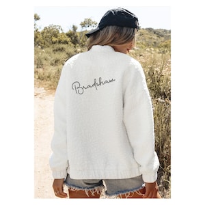 May include: White fluffy jacket with a black zip and a black cursive signature that says "Bradshaw" on the back.