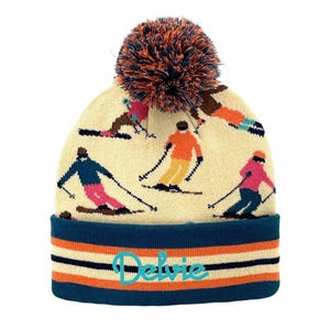 May include: Cream-colored knit beanie with a navy blue and orange striped brim. The hat features a pom-pom and a pattern of skiers in various colors. The name "Delvie" is embroidered on the brim.