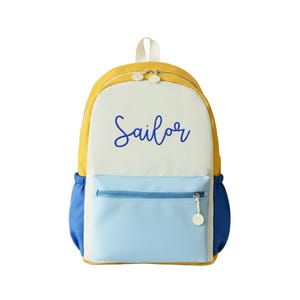 May include: A mustard yellow, cream, and light blue backpack featuring the word Sailor embroidered in blue script on the front panel. The bag includes a front zippered pocket with a circular pull tab and two side pockets with elasticized openings.