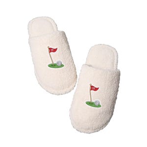 May include: A pair of white fuzzy slippers with a golf course embroidered on each slipper. The golf course design includes a green putting green, a golf ball, and a red flag with the number 19 on it.