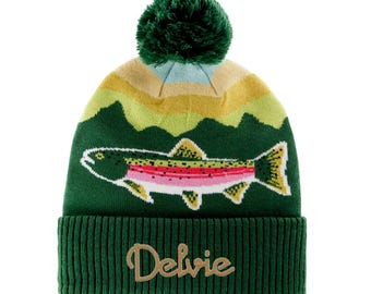Puede incluir: Gorro de punto verde con un gráfico de trucha colorida y un pompón verde oscuro. El gorro presenta la palabra "Delvie" en color dorado en la parte delantera. La parte superior del gorro tiene un diseño de montaña en tonos verdes, amarillos y beige.