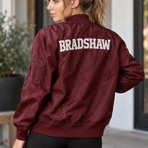 May include: A maroon bomber jacket featuring the name BRADSHAW embroidered in white block letters across the upper back. The jacket has a ribbed collar, cuffs, and hem, along with a utility pocket on the upper left sleeve with a zipper closure.